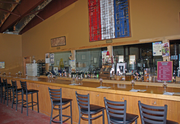 Visitors will find a large tasting room area with several antiques. Saddles make an interesting spot for visitors to have photos taken. There is plenty of room to spread out and there are tables and chairs in one section. The tasting counter is long and can accommodate a group. Wine enthusiasts sit at the tasting counter. Behind the tasting counter, windows provide a glimpse into the winery. Founders Wine Cellar